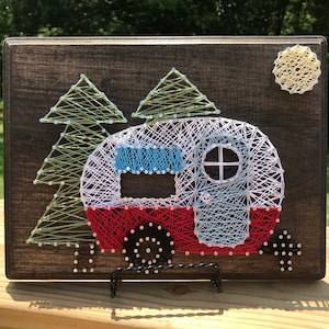 May include: String art of a red, white, and blue camper with a blue window and a yellow moon. The camper is surrounded by two green trees. The art is on a dark brown wooden background.