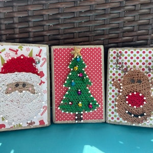 May include: Three string art Christmas ornaments. The first ornament is a Santa Claus with a white beard and red hat. The second ornament is a green Christmas tree with colorful ornaments. The third ornament is a brown reindeer with a red nose.