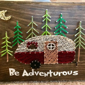 May include: String art of a red camper van with a white roof and a brown door, surrounded by green trees. The background is a dark brown wood. The text "Be Adventurous" is written below the camper.