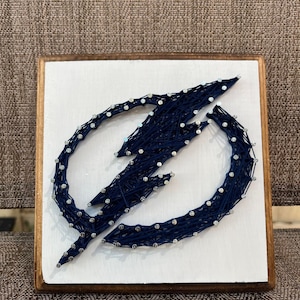 May include: A square string art piece featuring a navy blue lightning bolt design on a white background. The design is made with string wrapped around small nails, set in a wooden frame with a brown border.