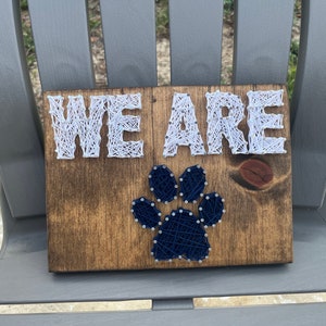 May include: A wooden sign with the words "WE ARE" in white string art and a blue paw print in the center.