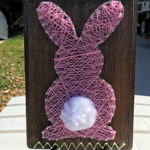 May include: Pink string art bunny on a wooden plaque with a white cotton tail. The bunny is outlined with green string.