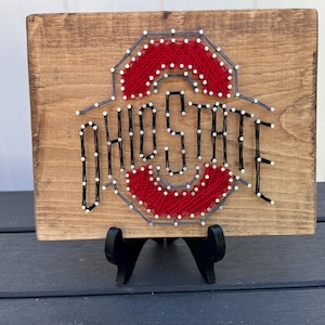 May include: Wooden sign featuring the Ohio State logo, crafted with red string and white dots on a brown wooden background. The word "OHIO STATE" is spelled out in black string. The sign is displayed on a black easel.