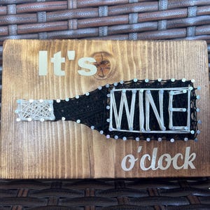 May include: A wooden sign with the text "It's Wine o'clock" in white letters. The word "Wine" is made with string wrapped around nails.