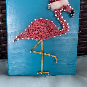 May include: A handmade flamingo string art piece on a teal blue wooden background. The flamingo is crafted with pink string and adorned with a red Santa hat with white trim. The flamingo's legs are made of gold string.