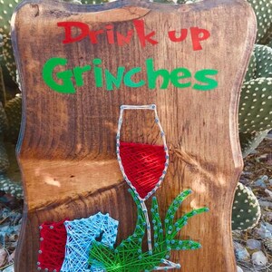 May include: Wooden sign with the text "Drink up Grinches" in red and green letters. A string art design of a wine glass with a green hand holding it is below the text.