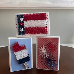 May include: Three wooden blocks with string art designs. The top block has a red, white, and blue American flag design. The middle block has a red, white, and blue popsicle design. The bottom block has a red, white, and blue firework design.