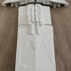 Puede incluir: Un modelo blanco impreso en 3D de una nave espacial, con un diseño detallado con múltiples secciones. El modelo está colocado sobre una superficie de madera. El diseño de la nave incluye múltiples torretas y un casco detallado.