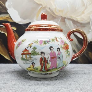 May include: A white porcelain teapot featuring a hand-painted scene of figures in traditional clothing near a pavilion. The teapot has a bright orange spout, handle, and lid rim, all accented with gold trim. Floral motifs surround the central scene.
