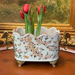 May include: A rectangular ceramic planter with a scalloped rim, featuring a floral and dragonfly pattern in pink, green, and brown. The vessel has ornate gold-toned metal handles shaped like butterflies and stands on four decorative metal feet.