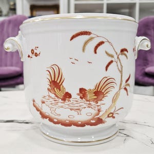 Op de afbeelding: Een witte keramische wijnkoeler met twee sierlijke handvatten en een gouden rand. De buitenkant is versierd met een roestkleurig patroon van twee hanen bij een gestileerde boom en kleine vogels die in de achtergrond vliegen.