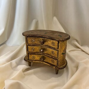 Pode incluir: Uma pequena cómoda em forma de rim com um acabamento dourado desgastado e padrões decorativos complexos em tons escuros. A peça inclui três gavetas curvas, cada uma com um puxador redondo, apoiada sobre quatro pernas curtas.