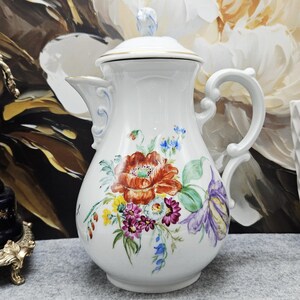 Puede incluir: Una cafetera de porcelana blanca con tapa, decorada con un arreglo floral pintado a mano de flores rojas, moradas, amarillas y azules con hojas verdes. La pieza presenta un asa curva, un pico decorativo y un borde dorado en la tapa.