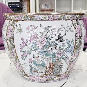 Pode incluir: Um vaso de cerâmica grande e redondo com base branca, apresentando ilustrações pintadas à mão de pavões coloridos, pássaros pequenos e flores cor-de-rosa e azuis. A borda é decorada com um padrão floral repetitivo em rosa e verde.