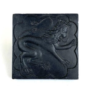 May include: A square black tile featuring a raised relief design of a lion in profile. The lion is depicted with a crown, an open mouth with a protruding tongue, and a long tail. The figure is enclosed within a scalloped, cloud-like border.