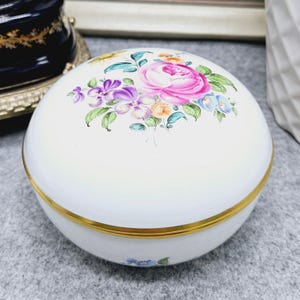 以下が含まれることがあります： 蓋に手描きの花柄が施された白い磁器製の丸い小物入れ。大きなピンクのバラ、紫のスミレ、緑の葉が描かれています。蓋と本体は金色の金属製の縁で繋がっています。