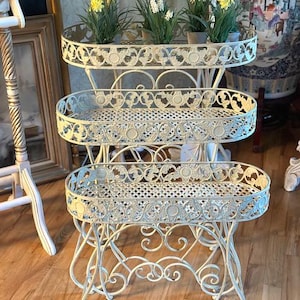 Pode incluir: Um suporte para plantas de metal em tom branco sujo com três níveis, apresentando prateleiras ovais com um padrão de treliça decorativo e pernas com volutas ornamentadas. A prateleira superior contém quatro pequenos vasos com flores amarelas e brancas.
