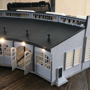 Puede incluir: Una estación de tren modelo gris con un techo curvo y múltiples ventanas. La estación tiene una pequeña puerta en el lateral y un letrero con un círculo y un número en la parte delantera.