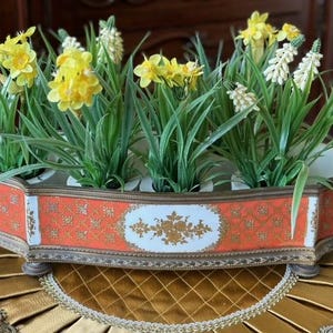 May include: An elongated, multi-opening ceramic flower trough featuring a vibrant orange and white exterior with intricate gold floral patterns and a metallic bronze-toned rim, filled with artificial yellow daffodils and white grape hyacinths.