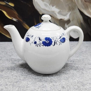 Könnte beinhalten: Eine weiße Teekanne aus Keramik mit rundem Körper, gebogenem Ausguss und passendem Deckel mit kleinem Knauf. Die Teekanne ist mit einem horizontalen Band aus komplexen blauen Blumenmustern und feinen schwarzen Rankenmotiven verziert.