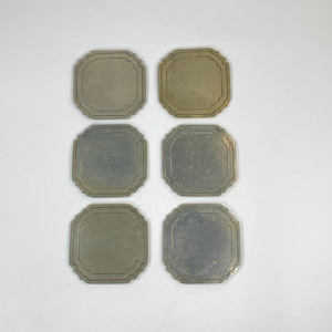 May include: A set of six square metal coasters with rounded corners and an octagonal inner border design. Each coaster features a smooth, flat surface with a muted, metallic finish, arranged in two vertical columns of three on a plain surface.