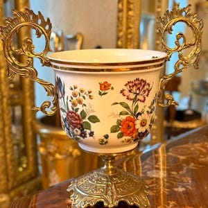 以下が含まれることがあります： 赤、オレンジ、青の花々が描かれたカラフルな花柄の白い磁器製壺。スクロールワークが施された2つの装飾的な金色の金属製ハンドルが付いており、複雑な彫刻が施された同色の金色の金属製台座の上に置かれています。