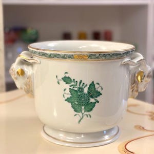 Pode incluir: Um cachepot de porcelana branca com um motivo floral verde na frente, uma borda decorativa verde e dourada ao longo da borda superior e duas alças esculpidas em forma de cabeça de carneiro com detalhes dourados nas laterais.