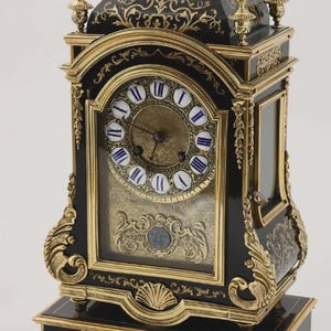 May include: An antique black and gold clock with ornate detailing. The clock features a round face with Roman numerals and blue accents. The clock has a rectangular shape with decorative gold accents and a base.