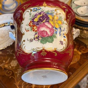 Op de afbeelding: Een dieprode keramische wijnkoeler met een centraal crèmekleurig paneel versierd met een levendig boeket van roze rozen, paarse pioenrozen en gele bloemen, geaccentueerd door gedetailleerd goudkleurig filigraan en twee handvatten aan de zijkanten.