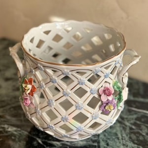 May include: A white porcelain basket with a lattice design, featuring gold trim along the rim and intersections. Small blue flowers are applied at each lattice crossing, with larger sculpted floral clusters in pink, orange, and purple on the sides.