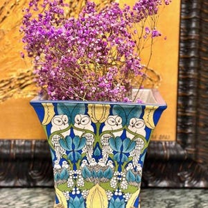 Op de afbeelding: Een vierkante keramische plantenbak met een herhalend patroon van witte uilen tussen gestileerd blauw en groen blad. De pot staat op vier sierlijke goudkleurige metalen pootjes en is gevuld met een boeket kleine, paarse gedroogde bloemen.