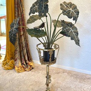 Pode incluir: Um suporte de latão para plantas com uma figura central de querubim sobre uma base circular, sustentando um aro metálico decorativo. Um vaso preto contém uma planta artificial Alocasia com folhas grandes, verde-escuras e em forma de seta.