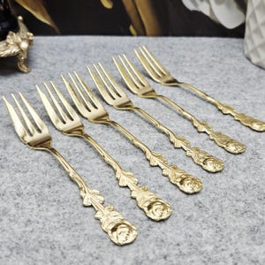 Op de afbeelding: Een set van vijf goudkleurige metalen vorken gerangschikt in een diagonale rij. Elke vork heeft een kop met vier tanden en een decoratief handvat met een gedetailleerd reliëf van een roos en bladeren over de gehele lengte.