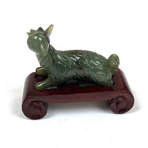 May include: A carved green stone figurine of a goat in a reclining position, featuring detailed fur textures and pointed horns. The animal sculpture is mounted on a dark reddish-brown wooden display stand with curved, scroll-like feet at each end.