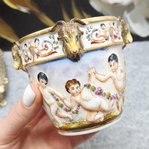 Może przedstawiać: Biały porcelanowy kubek z trójwymiarowymi akcentami w kształcie głów kozłów wzdłuż złoconego brzegu. Zewnętrzna strona ozdobiona jest ręcznie malowanymi scenami przedstawiającymi cherubiny trzymające girlandy kwiatowe na niebiesko-białym tle.