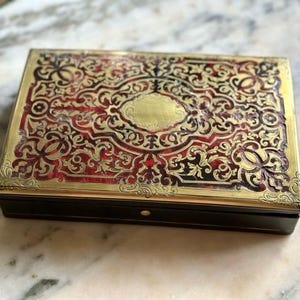 Pode incluir: Uma caixa retangular com uma base escura e uma tampa de metal dourado com padrões intrincados e elevados. A superfície metálica revela um fundo texturizado de cor vermelho profundo sob a filigrana dourada, com um oval central vazio.
