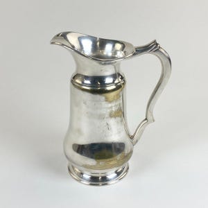 Puede incluir: Una jarra de metal plateado brillante con un pico vertedor curvo, un cuerpo cilíndrico alto con base redondeada y un asa decorativa. La superficie muestra áreas de brillo metálico y manchas visibles de tono dorado deslustrado.