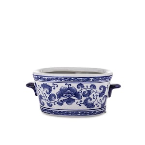 Pode incluir: Uma tigela de cerâmica branca com um padrão floral azul e duas alças laterais azul-escuras. O exterior apresenta um design repetitivo de flores estilizadas e vinhas, com bordas decorativas ao longo das extremidades superior e inferior.