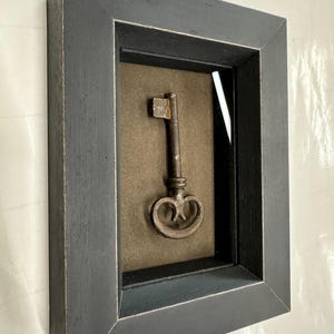 May include: A framed vintage key displayed in a shadow box. The key is a dark, aged metal with an ornate handle. The frame is a distressed black, adding to the antique aesthetic. The background is a muted brown.