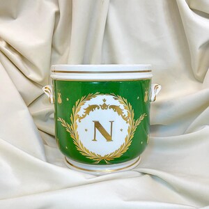 May include: A green ceramic vessel with two side handles, featuring a white central medallion with a gold letter N surrounded by a laurel wreath. The exterior is decorated with gold stars and bee motifs, with gold trim on the rim and base.