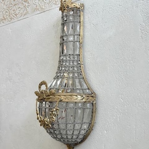 Puede incluir: Un aplique de pared con marco de metal dorado y detalles ornamentales en la parte superior e inferior. La lámpara está adornada con numerosos cristales de vidrio transparentes y rectangulares dispuestos en cuadrícula, unidos por un hilo plateado.