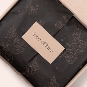 May include: A black gift box with a brown lid is open, revealing black tissue paper with a delicate pattern of butterflies and stars. The tissue paper has a small, light brown tag with the text "love of luna" printed in black.