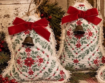 May include: Two bell-shaped fabric ornaments featuring a white background with a red and green poinsettia and holly leaf pattern. Each ornament has a frayed fabric edge, a dark red velvet bow, a metal bell, and a jute twine hanging loop.