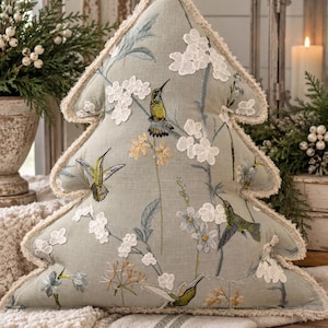 May include: A tree-shaped decorative pillow in sage green fabric featuring embroidered hummingbirds and white floral appliqués. The edges are finished with a frayed cream-colored trim, and the surface displays detailed botanical stitching.