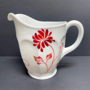 May include: White ceramic creamer pitcher with a red floral design. The pitcher has a curved spout, a handle, and a decorative flower and leaf pattern painted on the side. The creamer is a classic shape and design.
