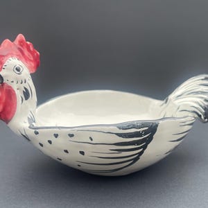 May include: A ceramic rooster-shaped bowl. The bowl is white with black accents and a red comb and wattle. The bowl is designed with a curved shape and is suitable for serving food.