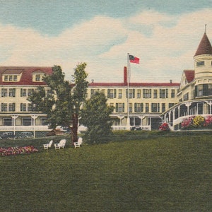 May include: Vintage postcard featuring a large, multi-story hotel with a light yellow facade and a red roof. The hotel has multiple windows and a porch. A flag flies in front of the building, and there are trees and chairs on the lawn.