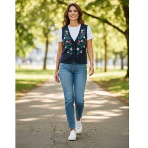 May include: A woman wearing a navy blue vest with floral embroidery over a white t-shirt and light blue jeans. The vest features pink and green floral designs. She is wearing white sneakers and walking on a paved path in a park.