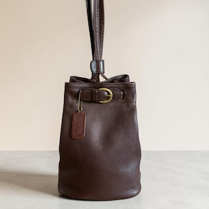 May include: A brown leather bucket bag with a drawstring closure and a gold-tone buckle. The bag has a long strap and a small leather tag attached with a gold chain. The bag is made of a smooth, textured leather.