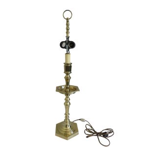 May include: A brass candlestick-style lamp with a hexagonal base and a ring at the top. The lamp features a small tray and a faux candle. A power cord with a plug extends from the base.